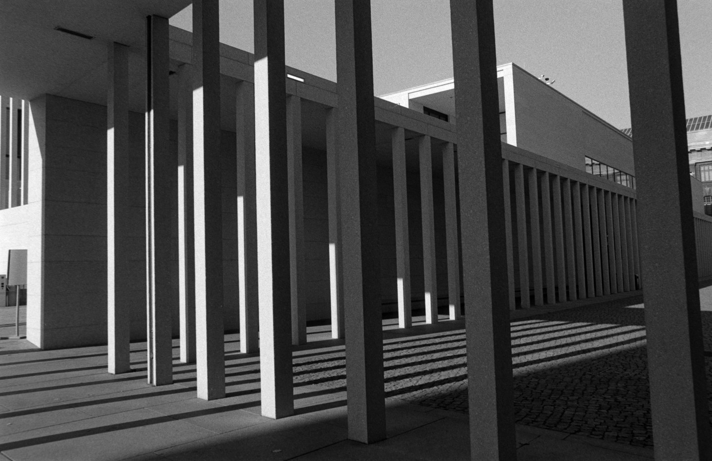 A series of tall, rectangular pillars cast long shadows on the ground in front of a modern building.