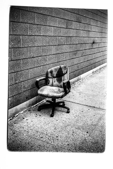 An old office chair sits on a sidewalk next to a brick wall.