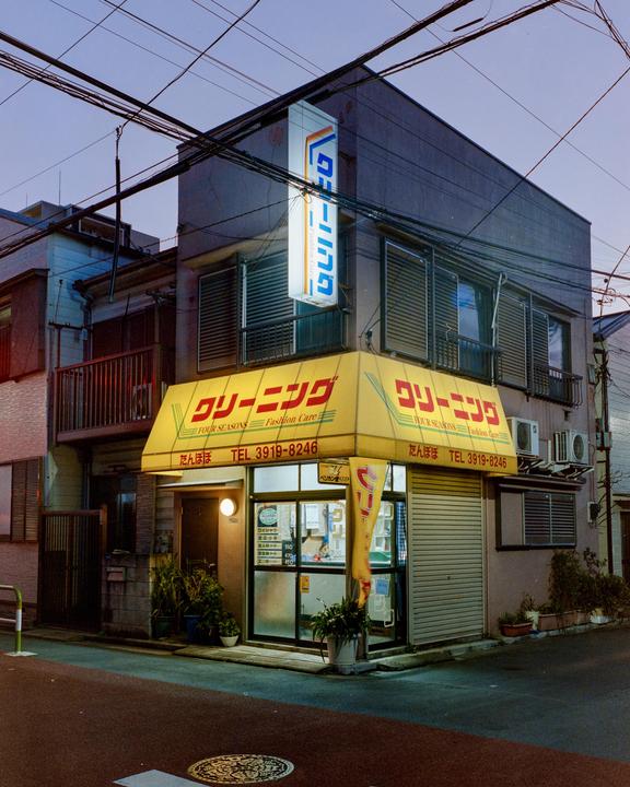 A brightly lit corner store with a yellow awning stands out on a dark street at dusk.