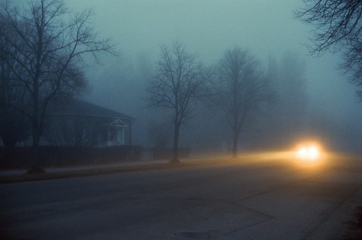 A car with bright headlights drives down a foggy street past a house and bare trees.