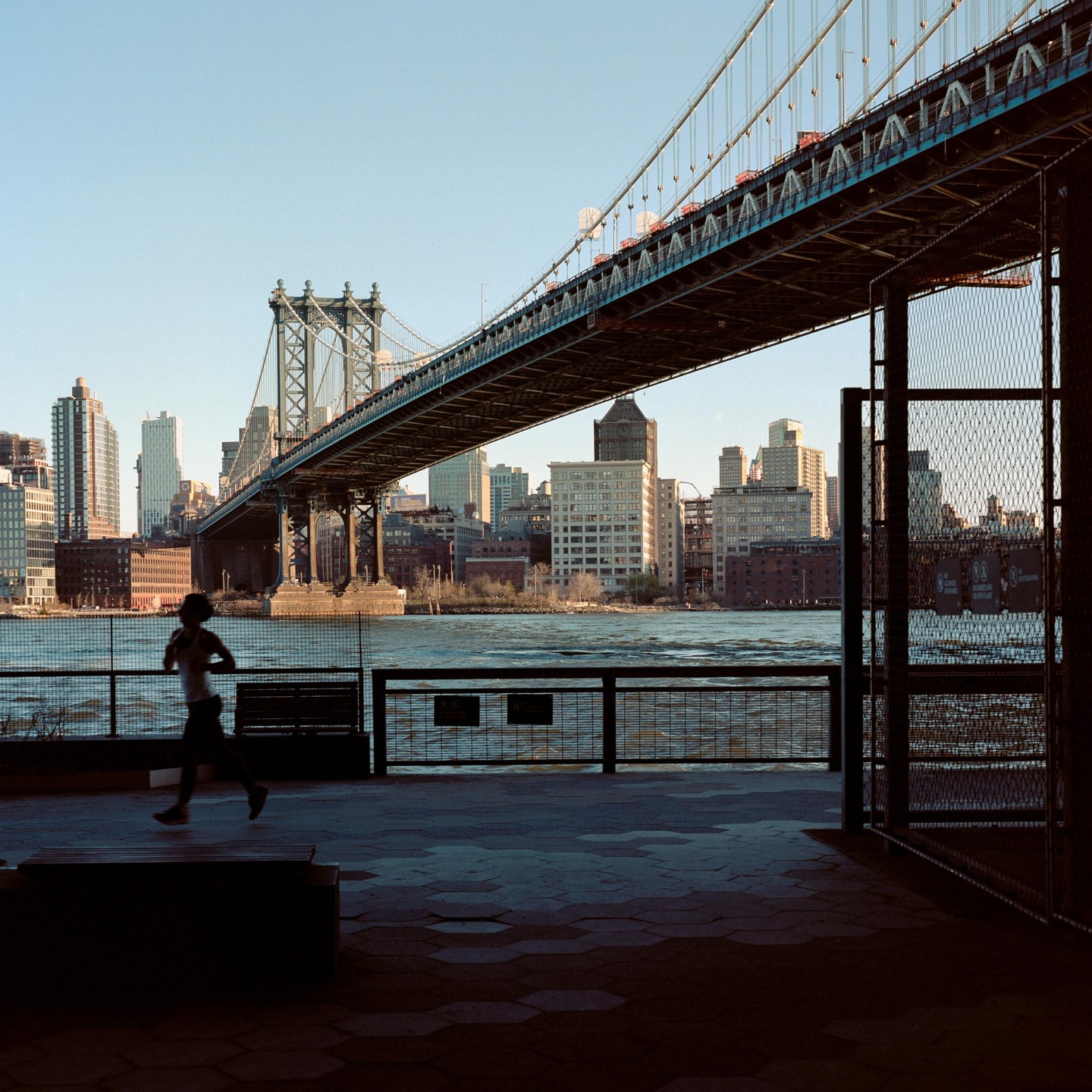 A person runs along a waterfront promenade with a large bridge and city skyline in the background.