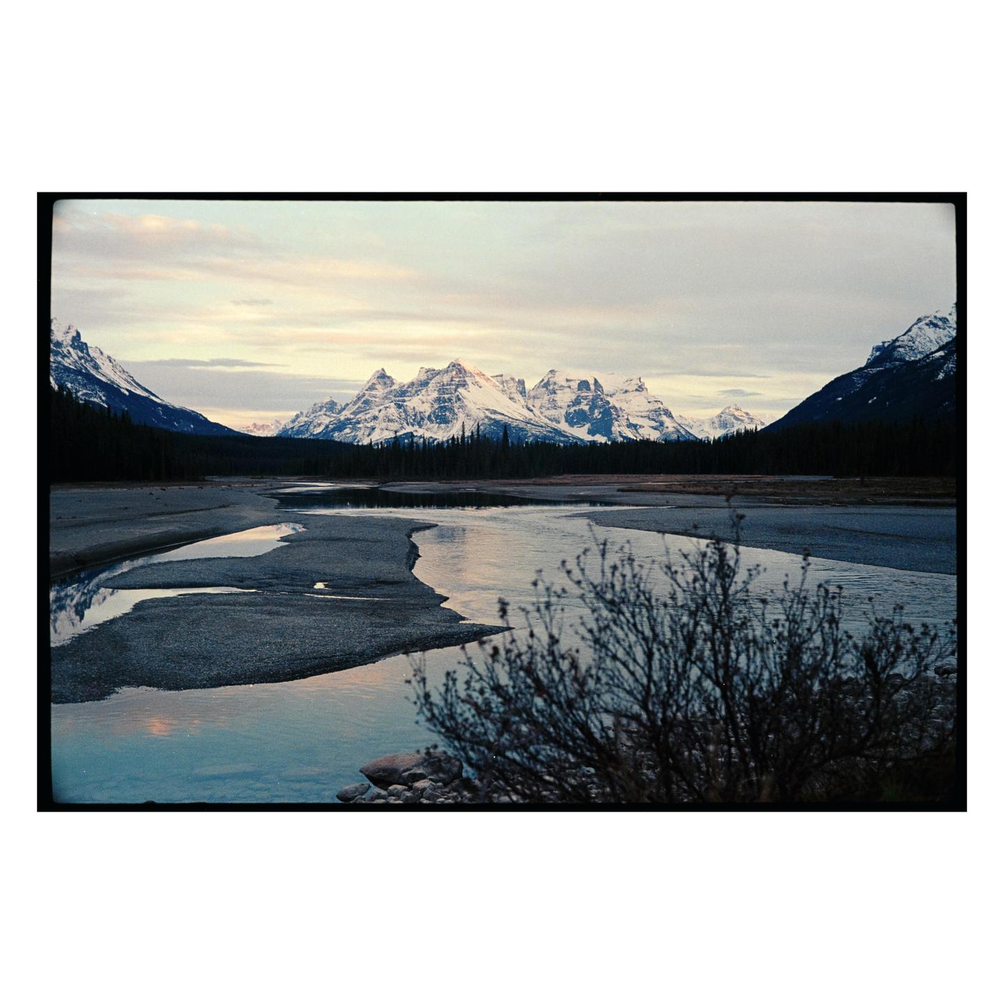 A river flows through a valley with snow-capped mountains and evergreen trees under a cloudy sky.