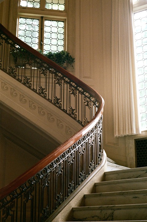 A grand staircase with an ornate railing and large windows is illuminated by natural light.
