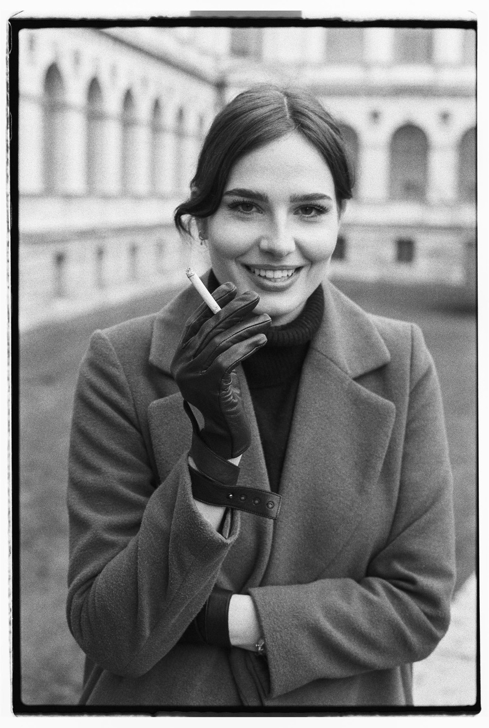 A woman in a coat and gloves smiles at the viewer while holding a lit cigarette.