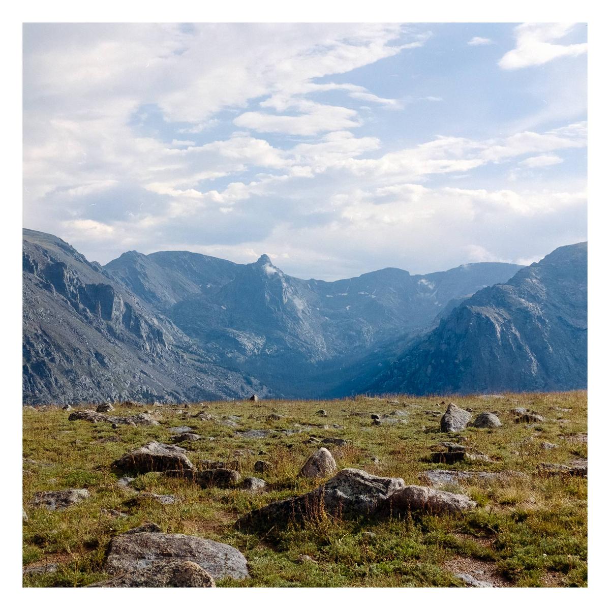A rocky, grassy field leads to a mountain range under a cloudy sky.