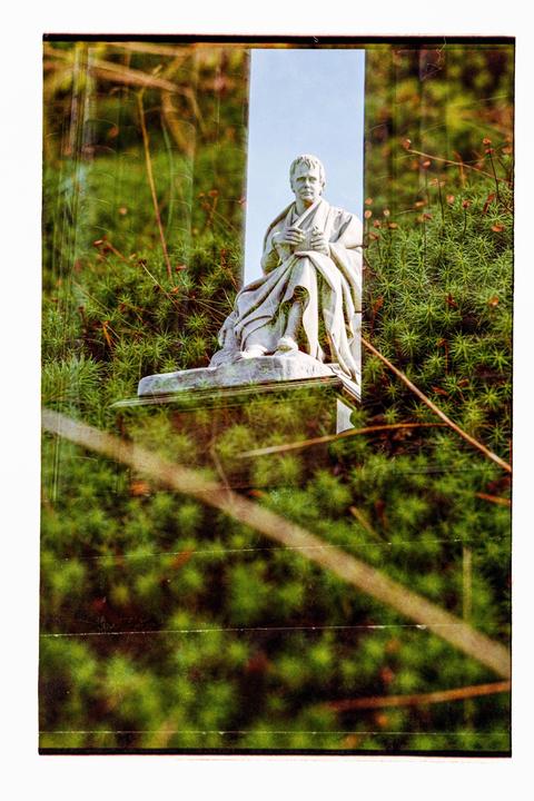 A statue of a man holding a book is visible through a double exposure with green foliage.