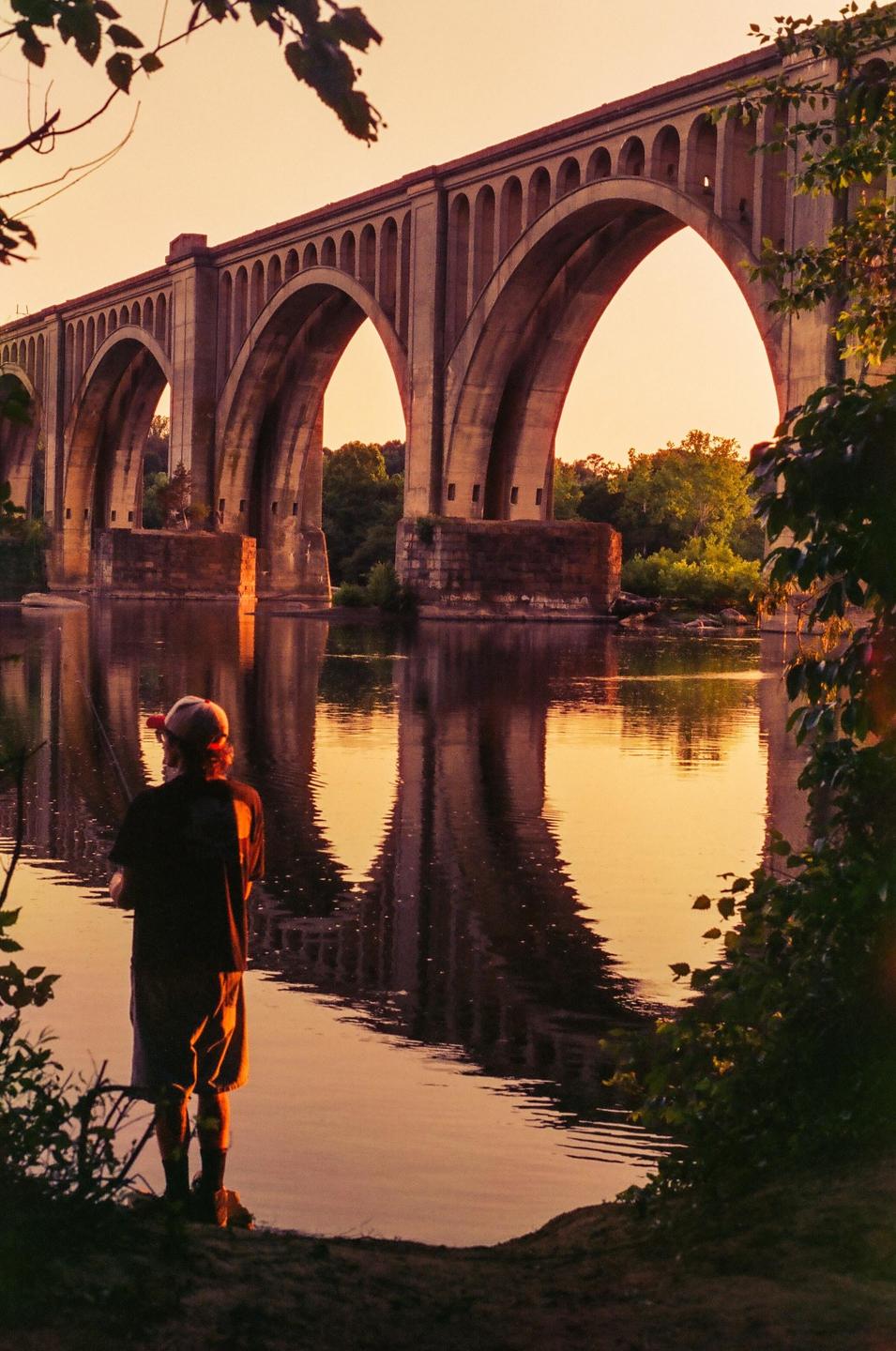 A man stands by a river, fishing, with a large arched bridge reflected in the water behind him at sunset.