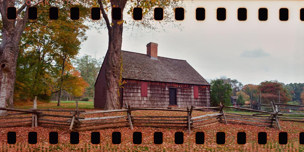 A rustic wooden house with a brick chimney stands behind a split rail fence, surrounded by trees with autumn foliage and fallen leaves.