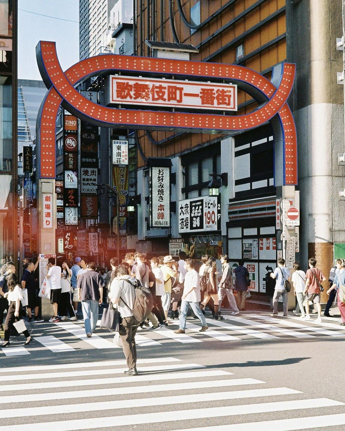 A crowd of people crosses a street in front of a large red archway and numerous signs on buildings.