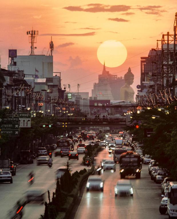 A large sun sets over a busy city street with cars and buildings, including a large Buddha statue and a temple.