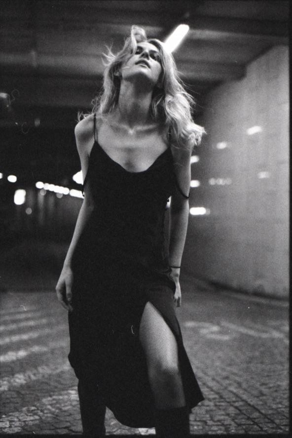A woman in a slip dress and fishnet stockings stands in a dimly lit underpass with her head tilted back.