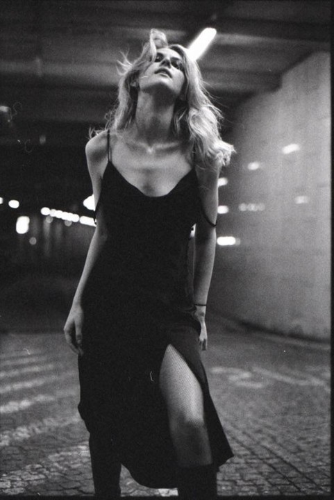 A woman in a slip dress and fishnet stockings stands in a dimly lit underpass with her head tilted back.