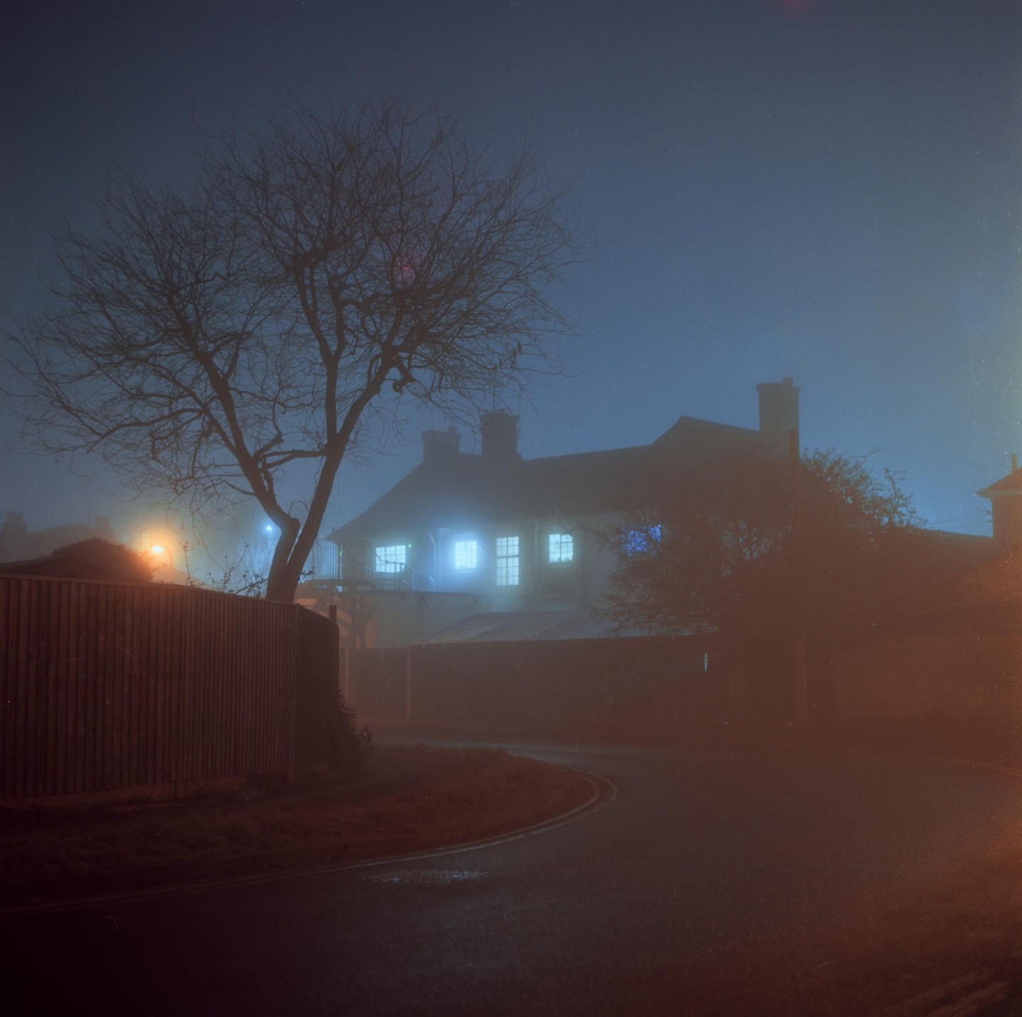 A house with glowing windows is visible through a thick fog at night, with a bare tree and a wooden fence in the foreground.