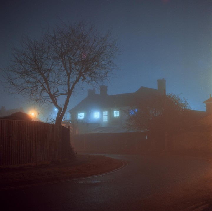 A house with glowing windows is visible through a thick fog at night, with a bare tree and a wooden fence in the foreground.