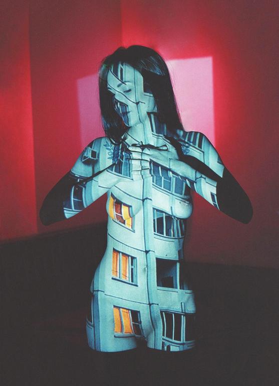 A woman with long hair has a building facade projected onto her body, with a red wall in the background.