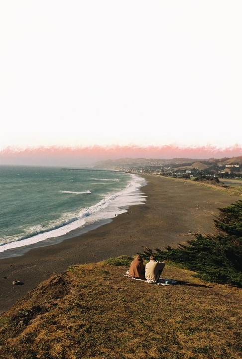 Two people sit on a grassy cliff overlooking a beach with waves crashing, a pier in the distance, and a town along the coast under a bright sky with a pinkish horizon.