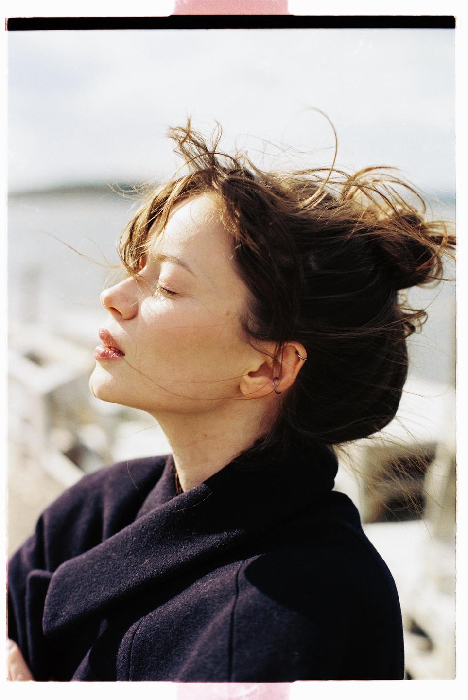 A woman with her eyes closed and hair blowing in the wind is illuminated by sunlight.