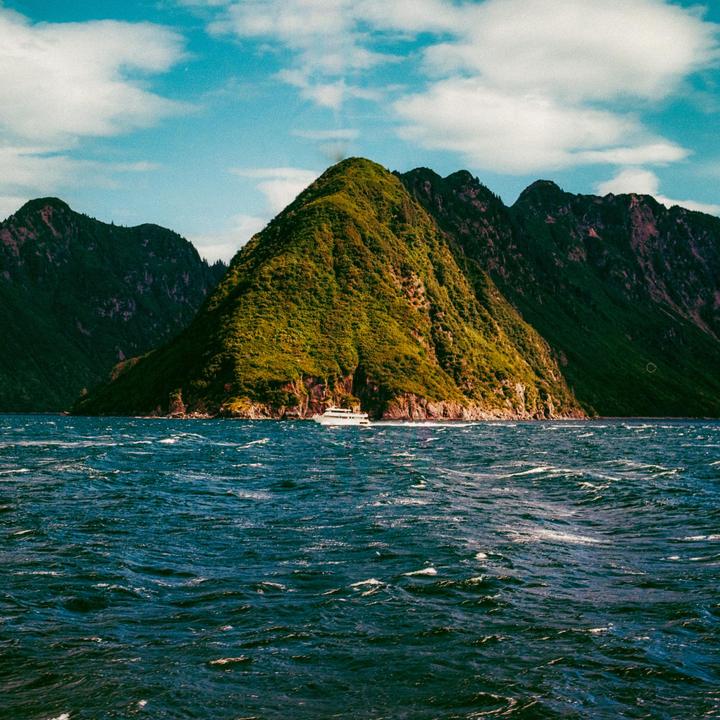 A white boat travels across choppy water with a large, green, conical mountain in the background under a blue sky with clouds.