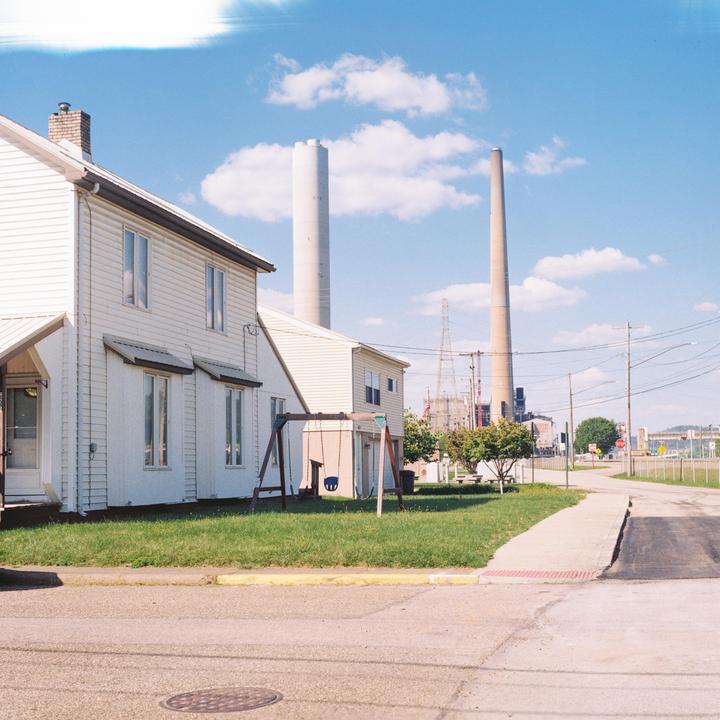 A residential street with houses and a swing set is shown with industrial smokestacks and power lines in the background under a blue sky with clouds.