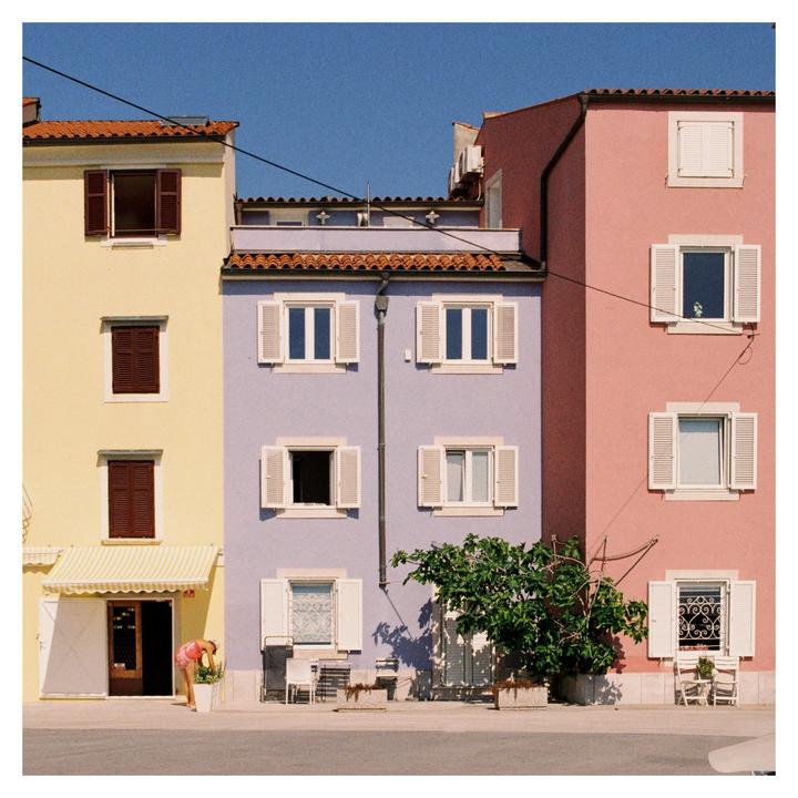 A woman tends to a plant in front of colorful buildings with white shutters under a clear blue sky.