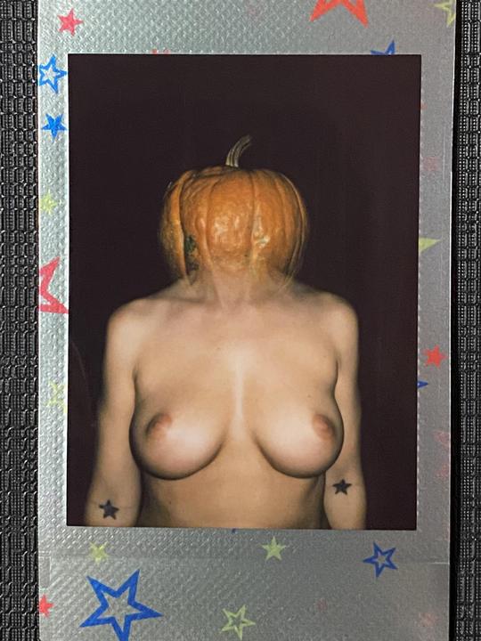 A person with a pumpkin on their head stands topless against a dark background.