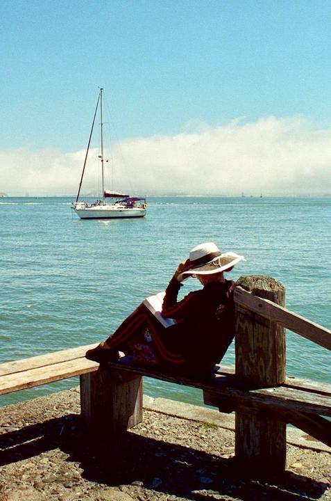 A woman in a hat sits on a wooden bench by the water, reading a book, with a sailboat in the background.