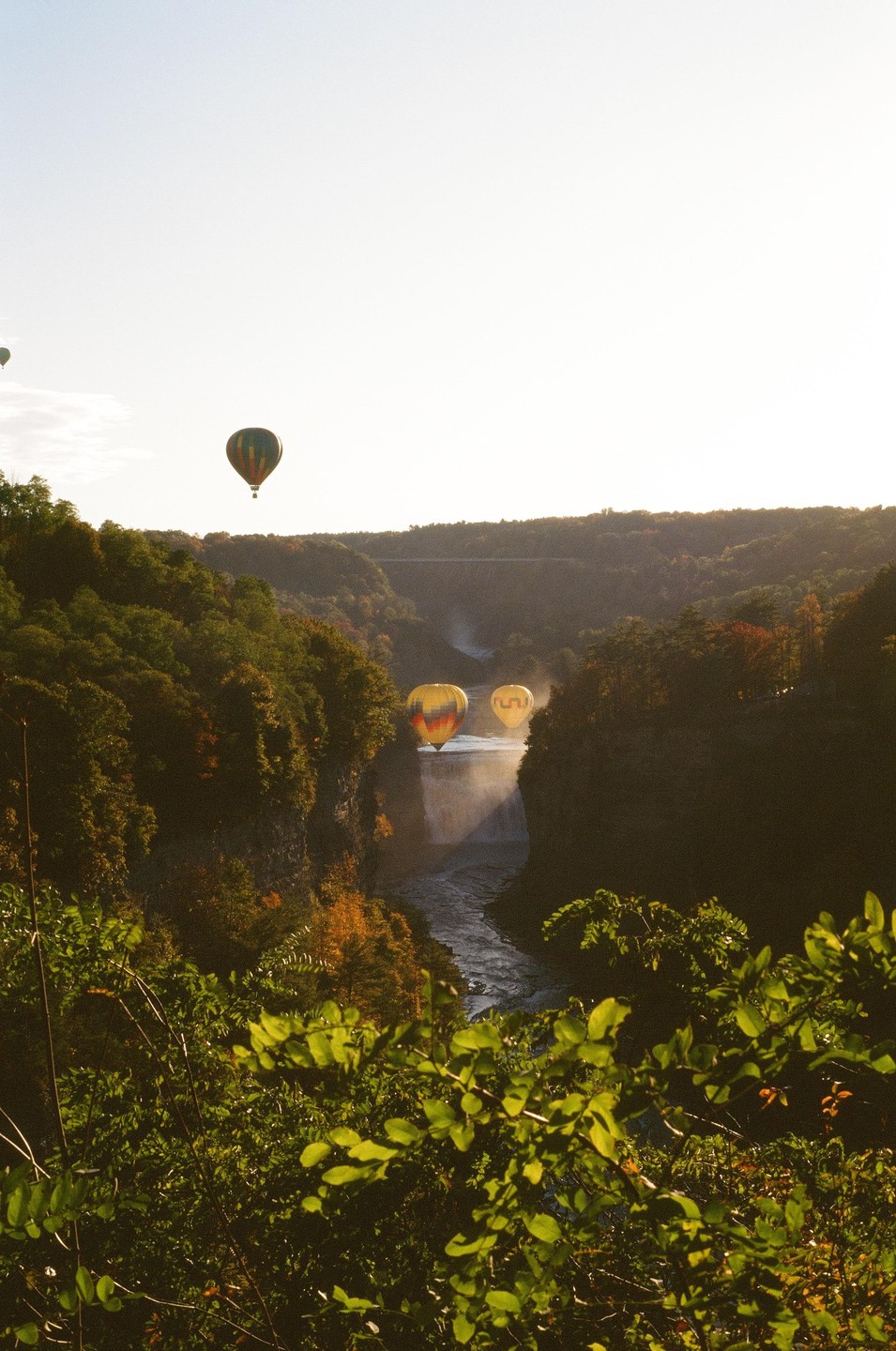 Three hot air balloons float above a river and waterfall surrounded by autumn foliage.