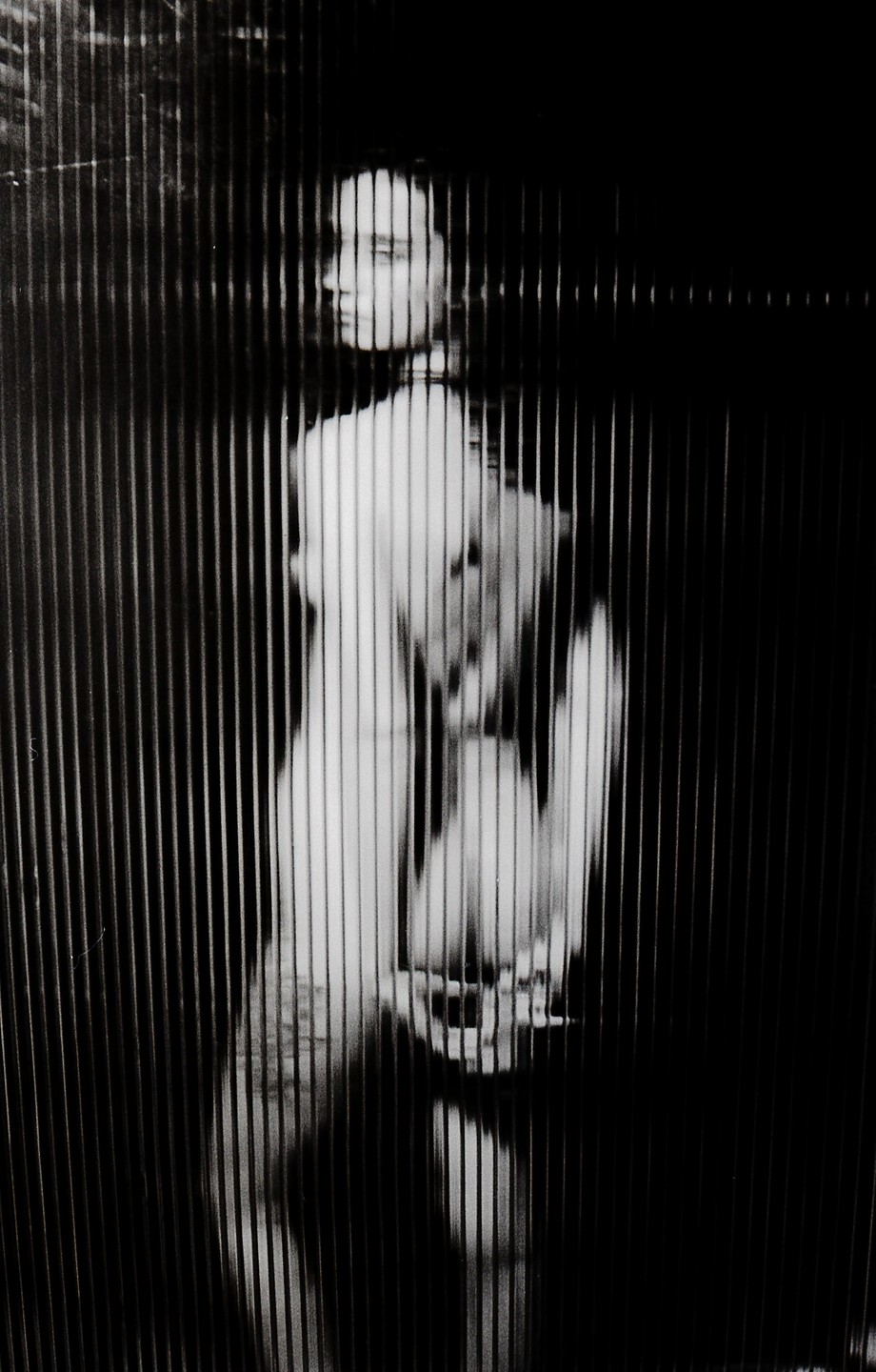 A woman is visible through vertical blinds in a black and white image.