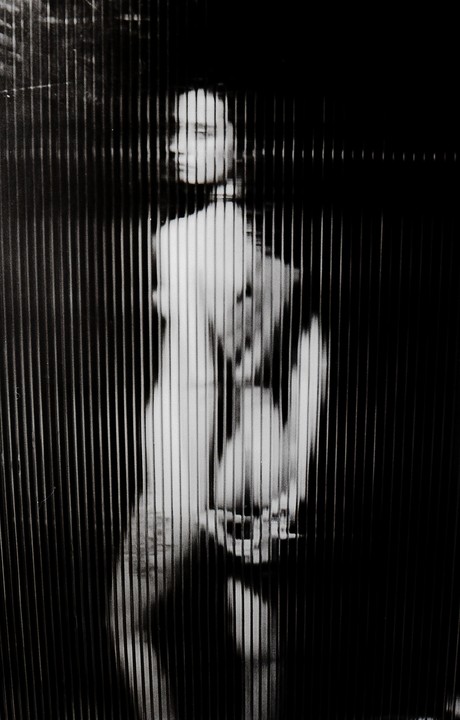 A woman is visible through vertical blinds in a black and white image.