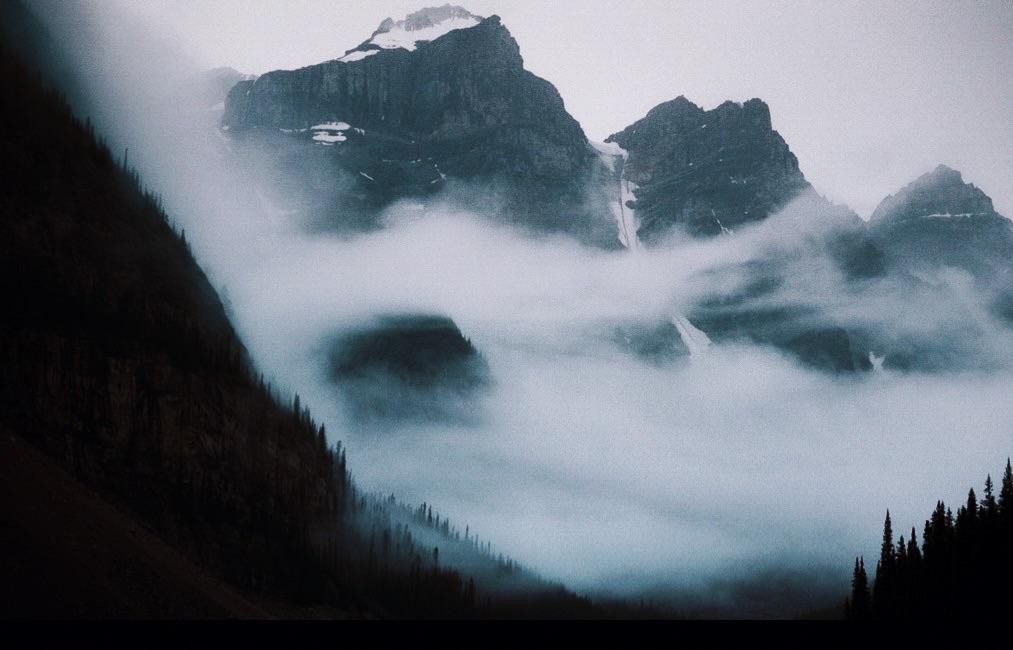 Fog rolls through a valley with mountains and pine trees on either side.