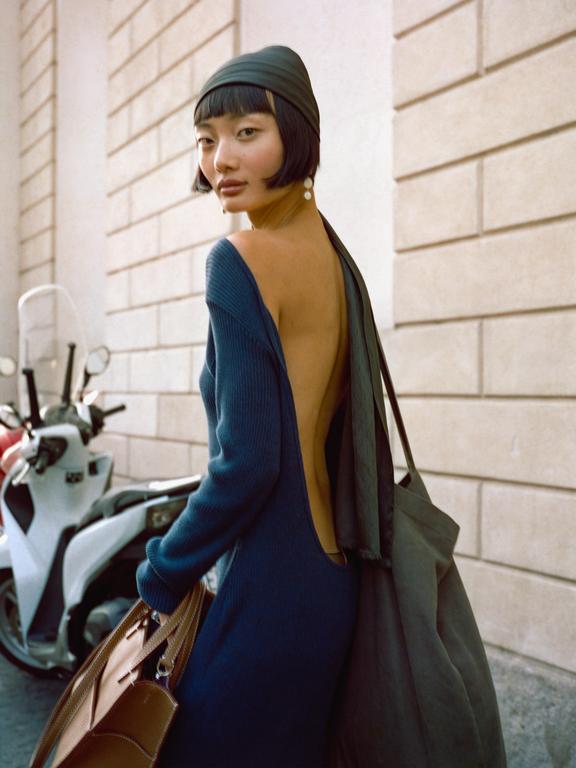 A woman in a blue dress with an open back looks over her shoulder while standing next to a scooter.