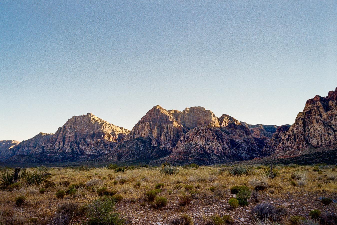 A desert landscape shows mountains in the background and dry brush in the foreground under a clear sky.