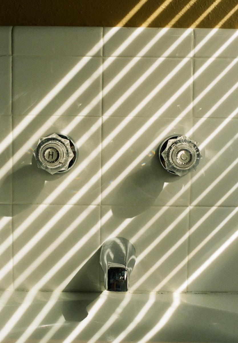 Sunlight streams through a window, casting striped shadows across a tiled bathroom wall with two faucet handles and a spout.