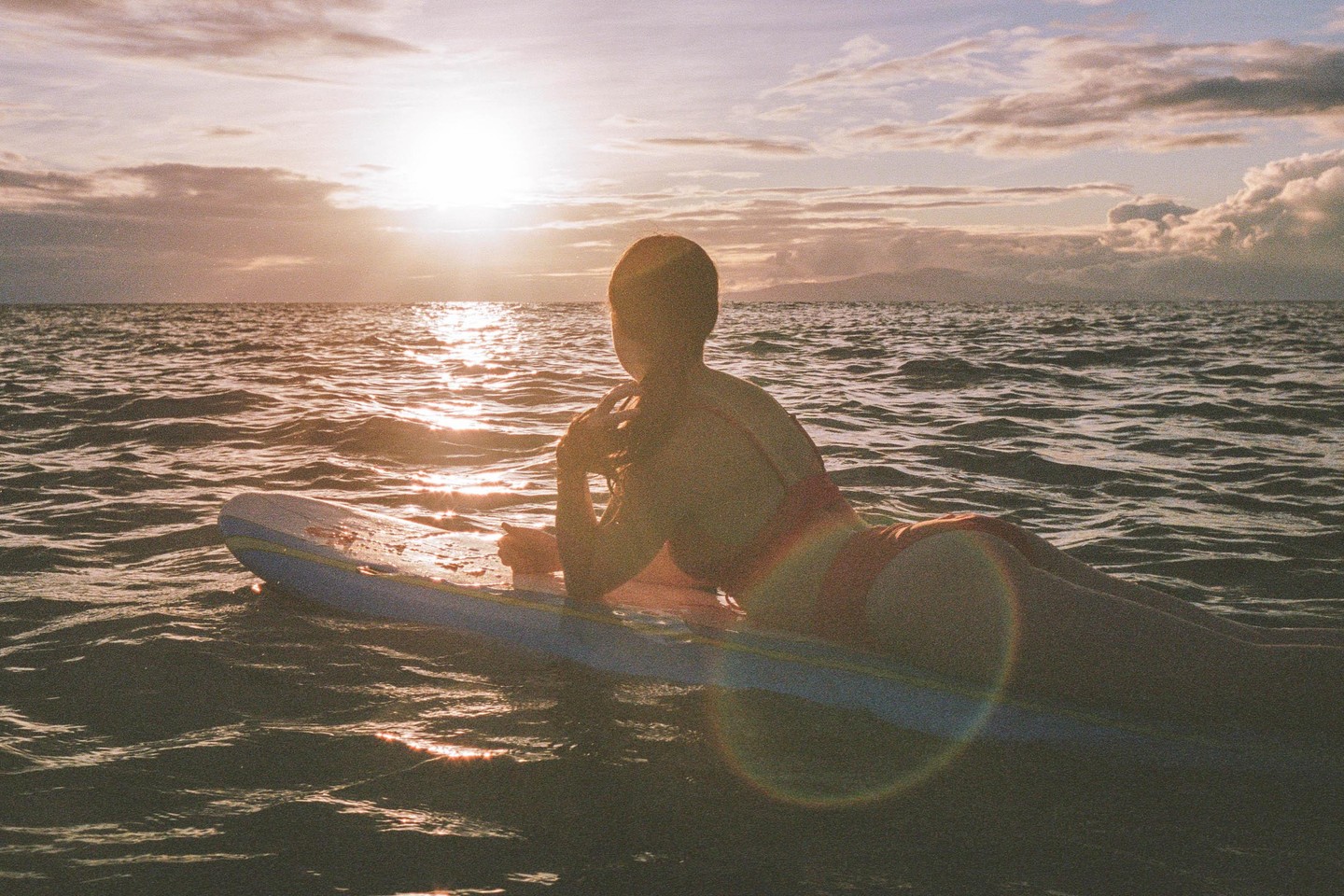A woman in a swimsuit lies on a surfboard in the ocean at sunset.