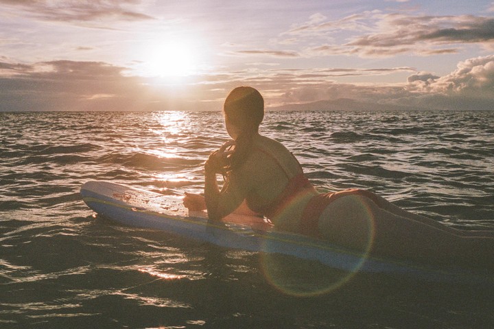 A woman in a swimsuit lies on a surfboard in the ocean at sunset.