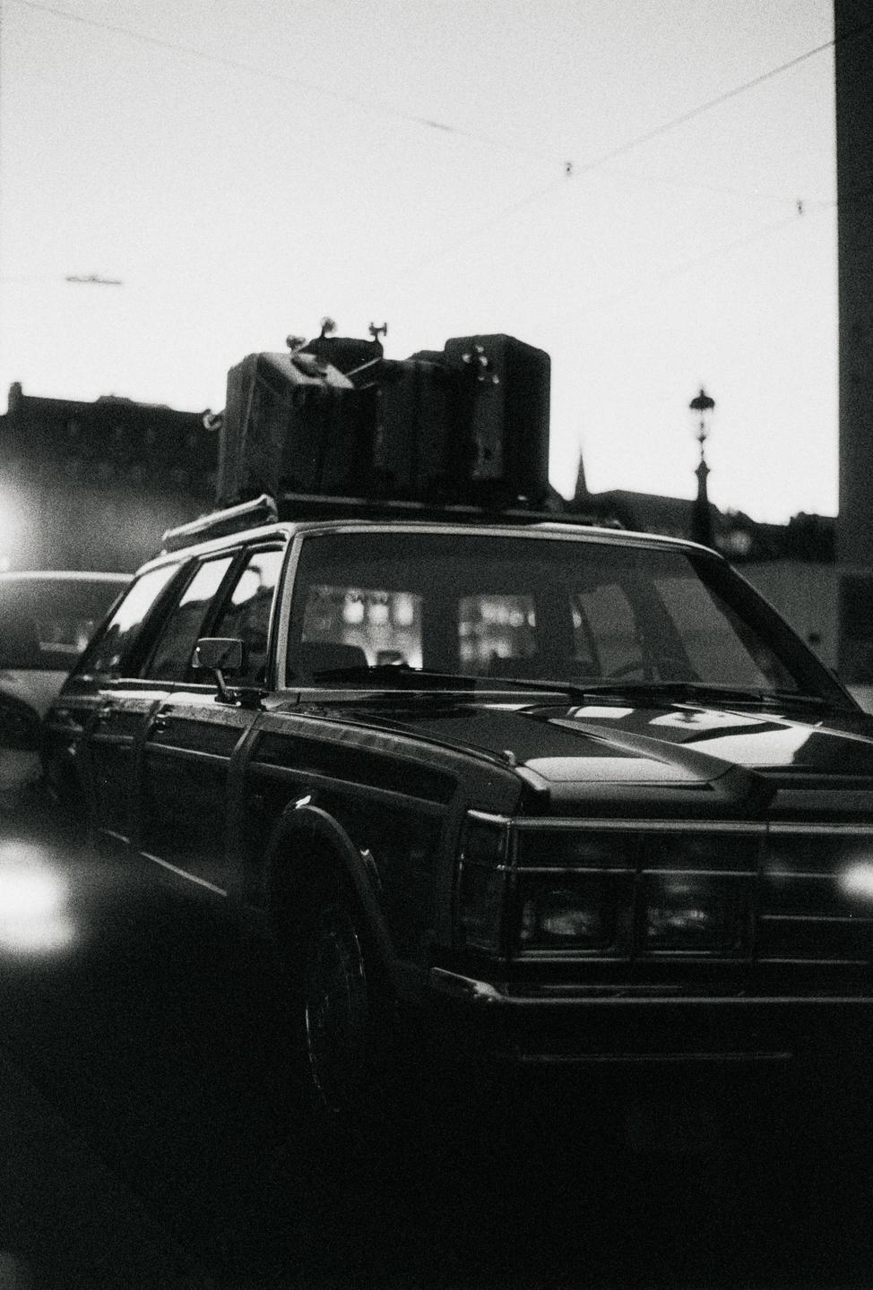 A car with luggage on its roof rack is parked on a street at dusk.