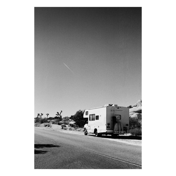 A recreational vehicle is parked on the side of a desert road with a contrail visible in the sky.