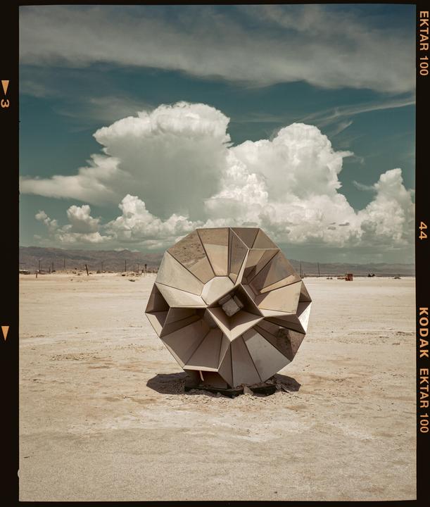 A geometric sculpture sits in a vast desert landscape under a cloudy sky.