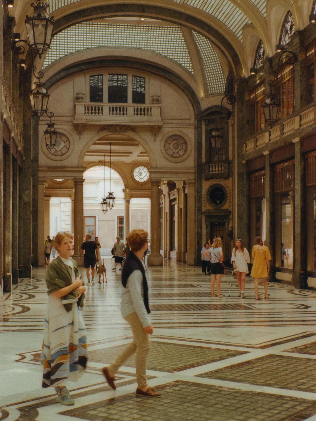 People walk through a grand, ornate hall with arched ceilings and decorative lamps.