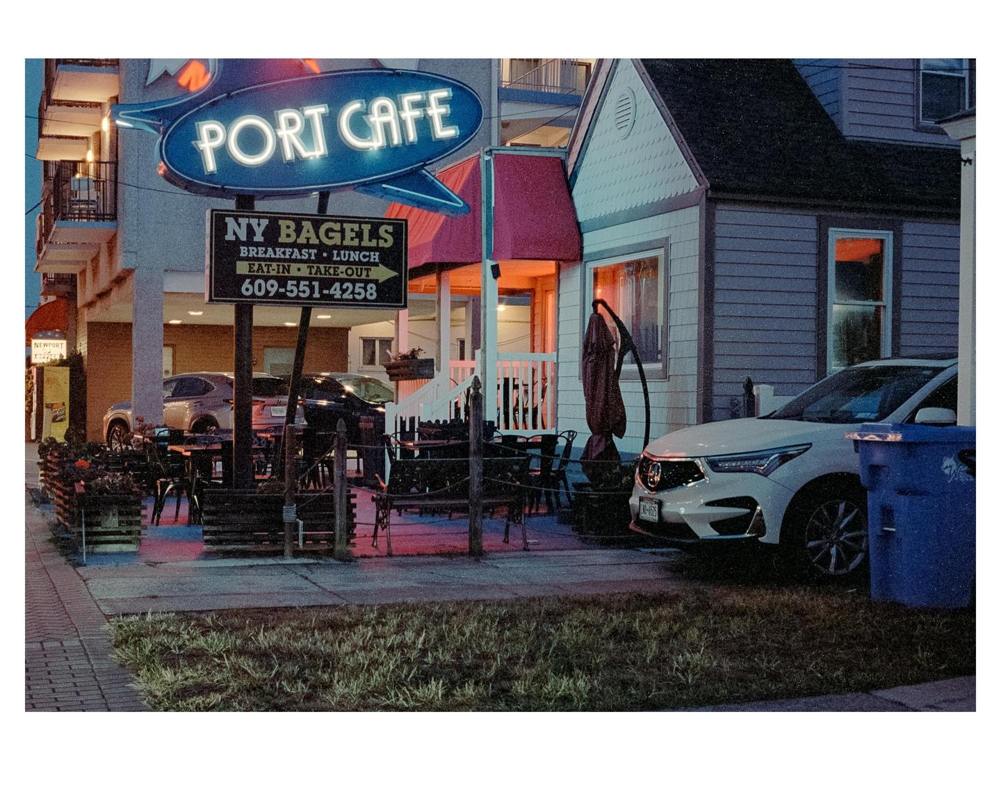A neon sign for Port Cafe and NY Bagels is illuminated above an outdoor seating area next to a building and parked car.