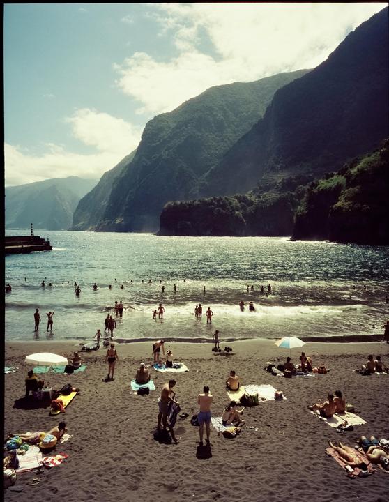 A beach with many people swimming and sunbathing is nestled between a calm ocean and large, verdant mountains.