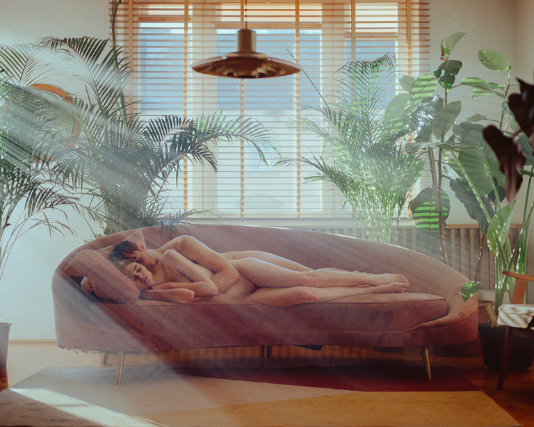 A nude couple lies on a pink sofa surrounded by large green plants, with sunlight streaming through a window.