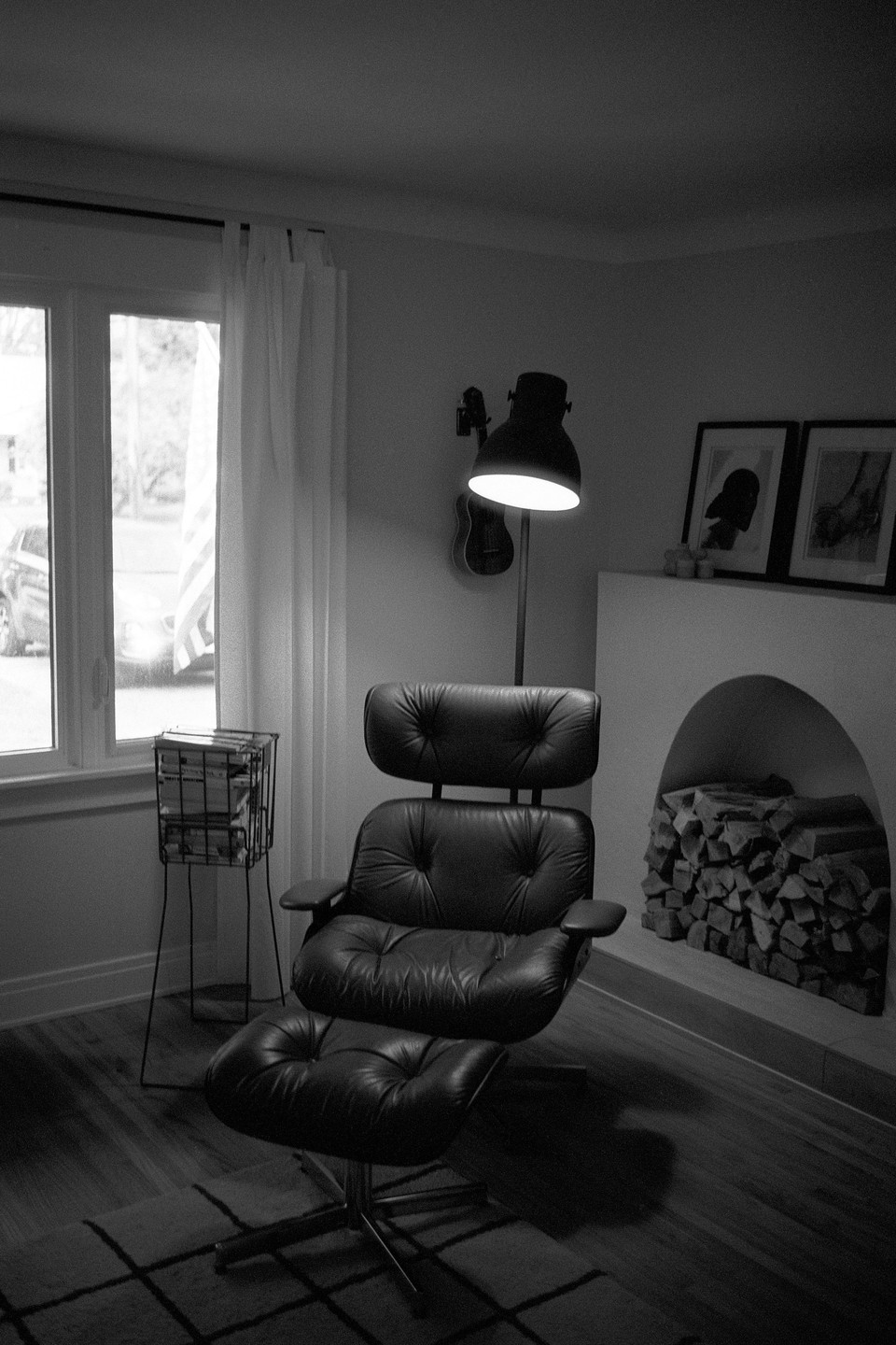 A black leather lounge chair and ottoman sit in a living room next to a window, a fireplace with stacked wood, and a floor lamp.