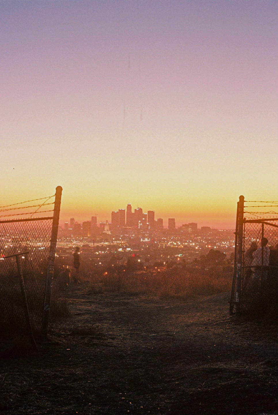 A city skyline is visible in the distance at dusk, framed by two fence posts and barbed wire in the foreground.