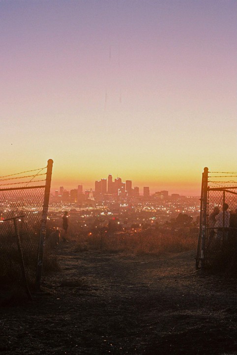 A city skyline is visible in the distance at dusk, framed by two fence posts and barbed wire in the foreground.