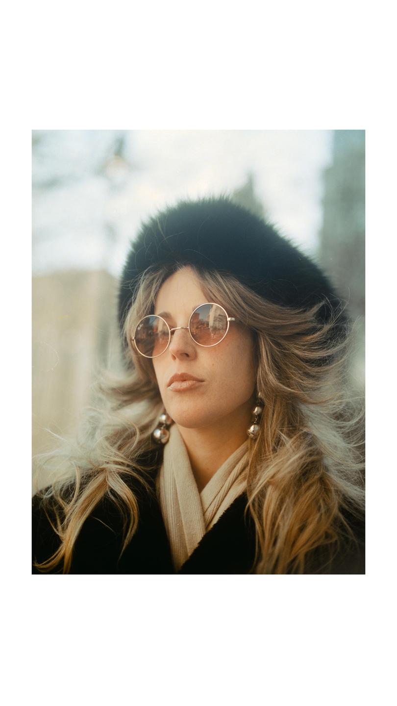 A woman with long blonde hair, round sunglasses, and a fur hat looks to the side.