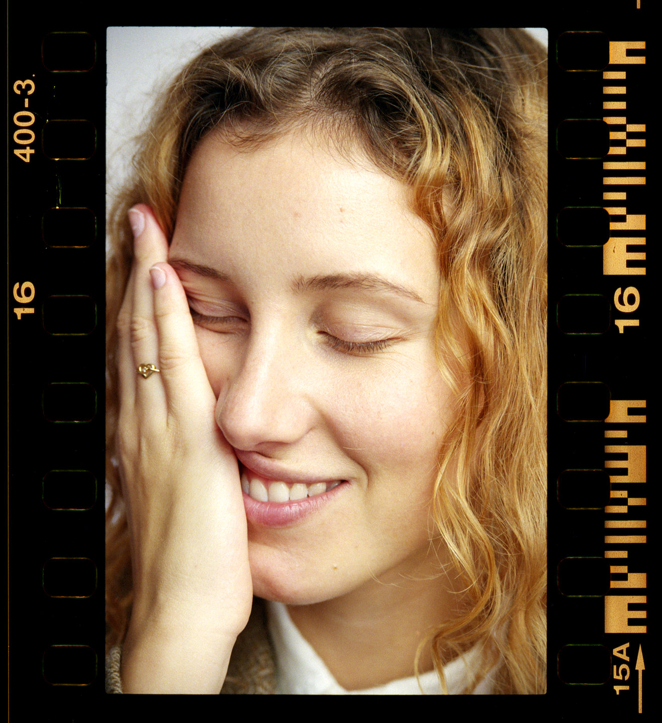 A woman with curly hair smiles with her eyes closed, her hand touching her cheek.