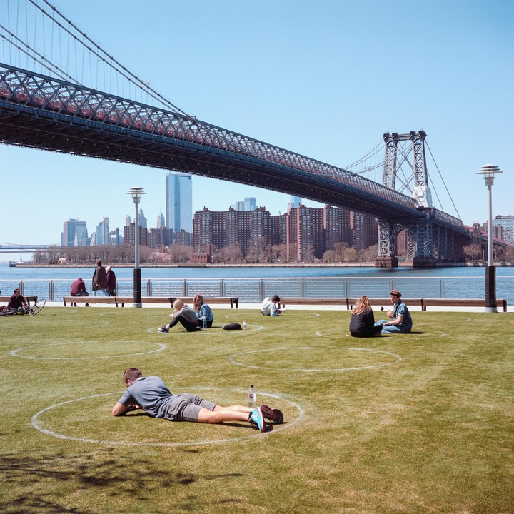 People relax on a sunny day in a park with a bridge and city skyline in the background.