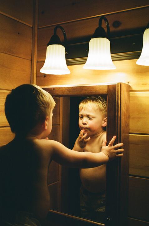 A young boy looks at his reflection in a mirror under two bright lights.