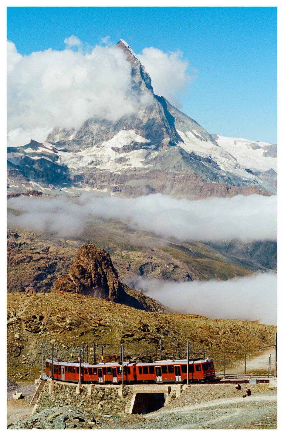 A red train travels along a track in a mountainous landscape with clouds partially obscuring a snow-capped peak.